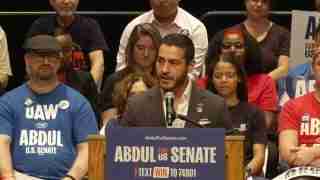 Man in a suit speaks at a campaign rally behind a podium that reads 'Abdul for US Senate'; crowd seated in blue and red shirts surrounds him.