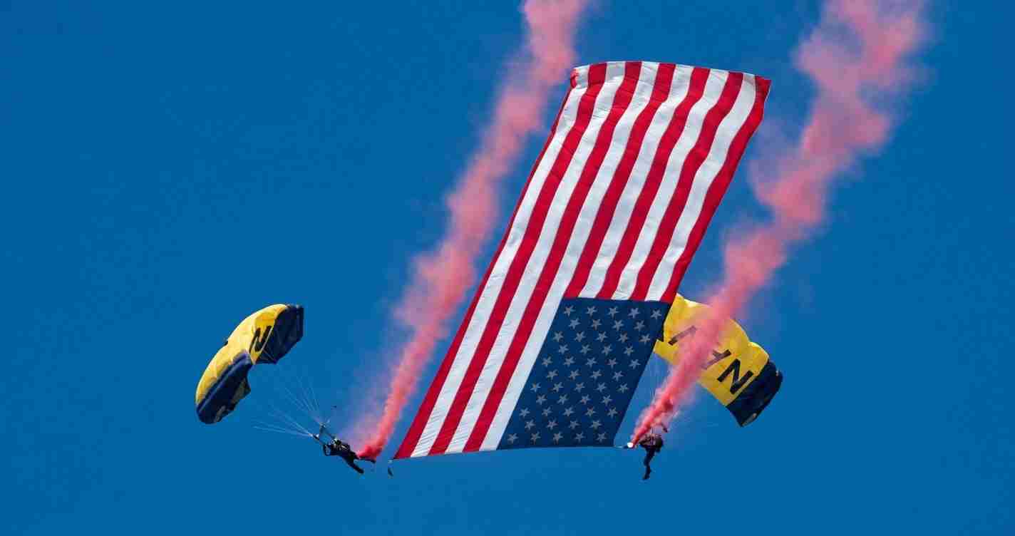 Two skydivers deploy a flag-shaped parachute resembling the U.S. flag, with red and white stripes and a blue field of stars, pink smoke trails behind the jumpers.