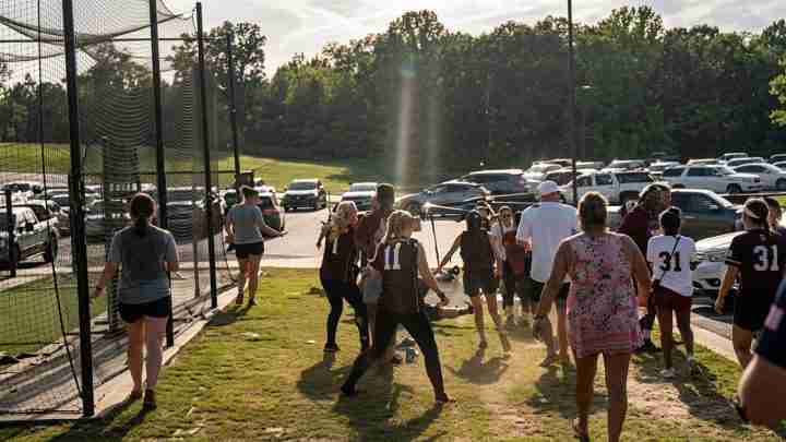 Group of people celebrating and playing on a sunny grassy field beside a tall chain-link fence, with cars in the parking lot behind them.