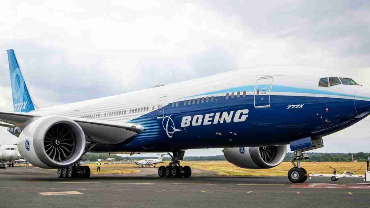 Side view of a large Boeing passenger jet (blue and white livery) parked on the tarmac with ground crew nearby and engines visible to the left and right.