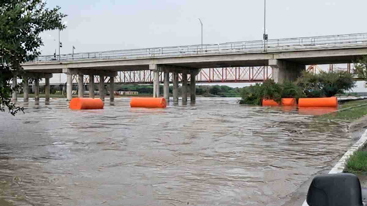 Flooded roadway underneath a bridge with orange floating barriers and submerged vegetation.