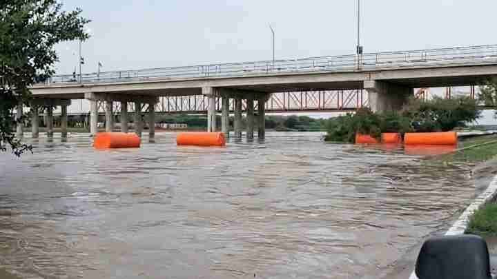 Flooded roadway underneath a bridge with orange floating barriers and submerged vegetation.