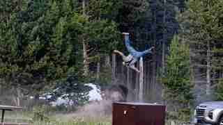 Man performing a backflip mid-air off a dirt ramp in a forested parking area, kicking up dust with a two-car background and trees nearby.