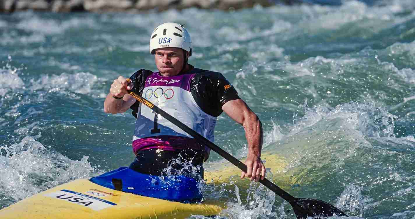 Athlete in a white helmet paddles a yellow kayak through choppy water with Olympic rings on the vest and 'USA' on the bow badge.
