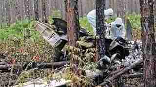 Two investigators in white hazmat suits examine a crashed aircraft fuselage tangled in fallen trees in a pine forest