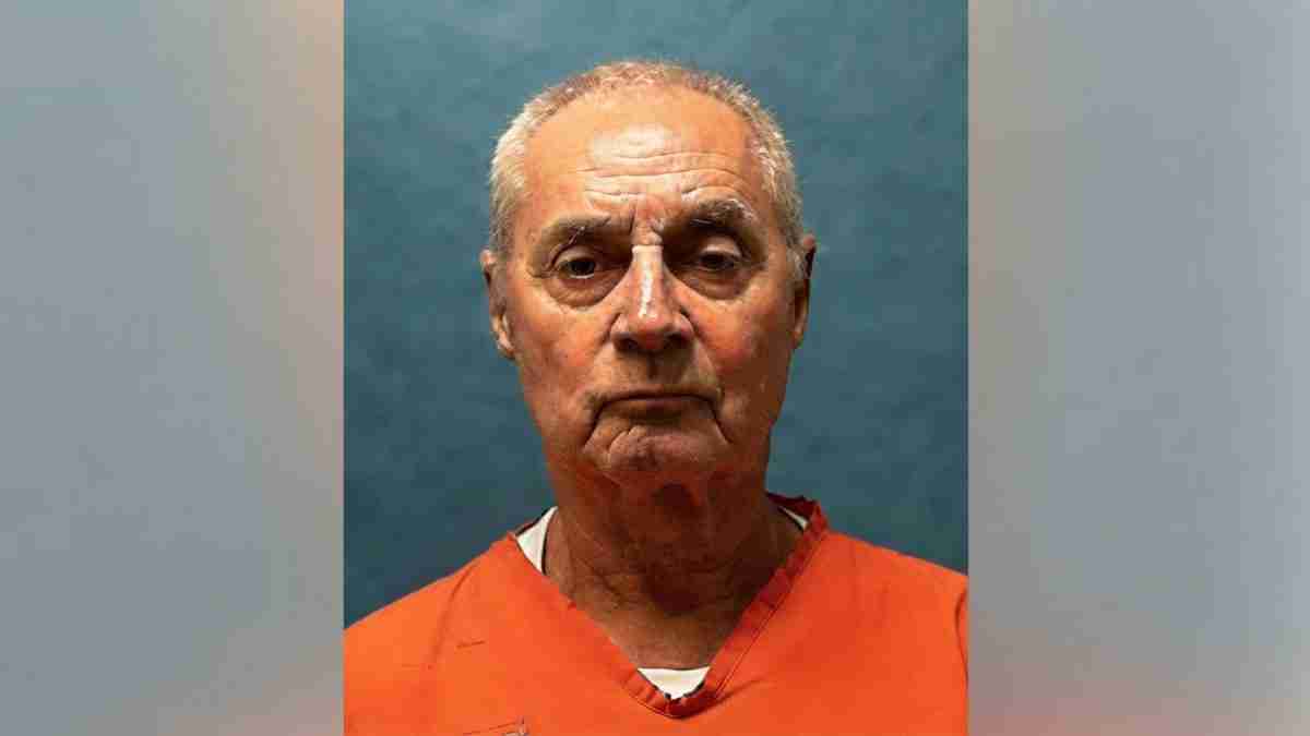 Mugshot of an older man wearing an orange prison jumpsuit against a blue backdrop, facing the camera.