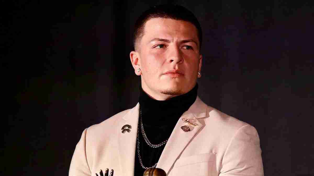 Portrait of a man in a beige blazer with a black turtleneck and silver chains, looking to the right against a dark backdrop.