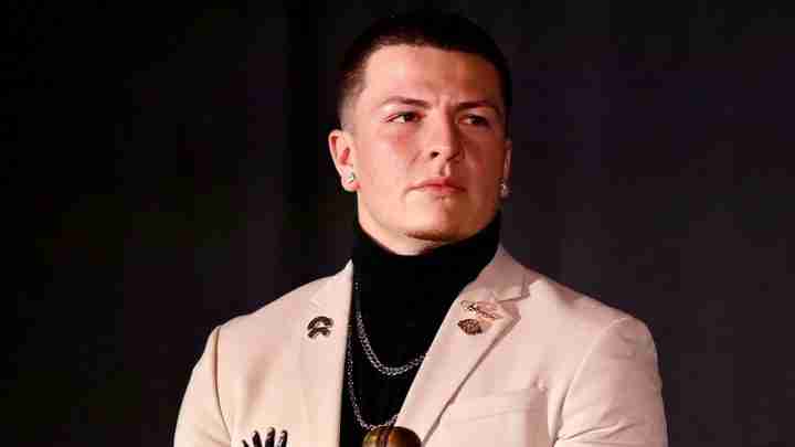 Portrait of a man in a beige blazer with a black turtleneck and silver chains, looking to the right against a dark backdrop.