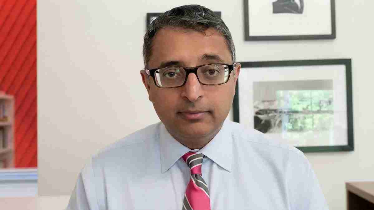 Man wearing glasses and a light blue shirt with a pink-striped tie sits in an office, looking at the camera.
