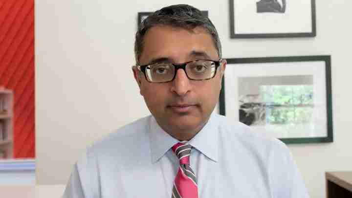 Man wearing glasses and a light blue shirt with a pink-striped tie sits in an office, looking at the camera.