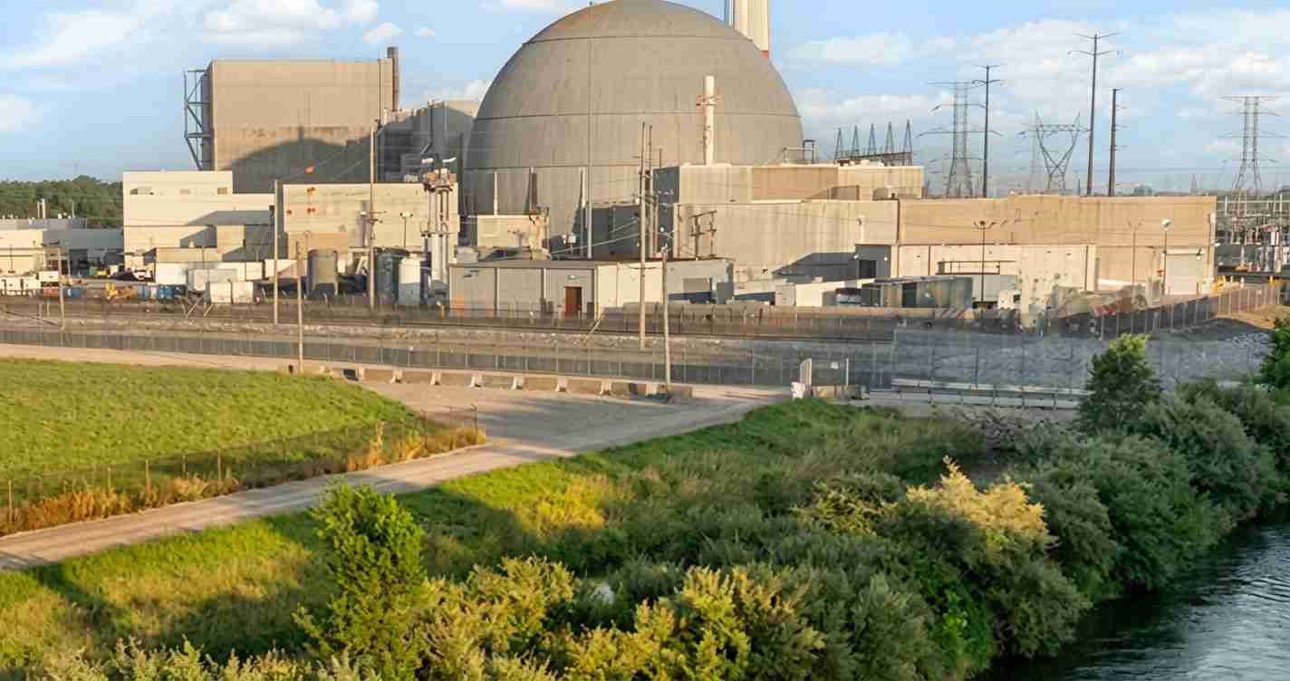 Nuclear power plant with a large domed reactor building behind a security fence, beside a grassy riverbank.