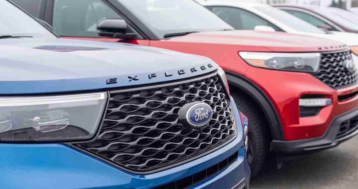 Close-up of a blue Ford Explorer front grille with the Ford emblem, parked alongside a red SUV at a dealership lot of cars.