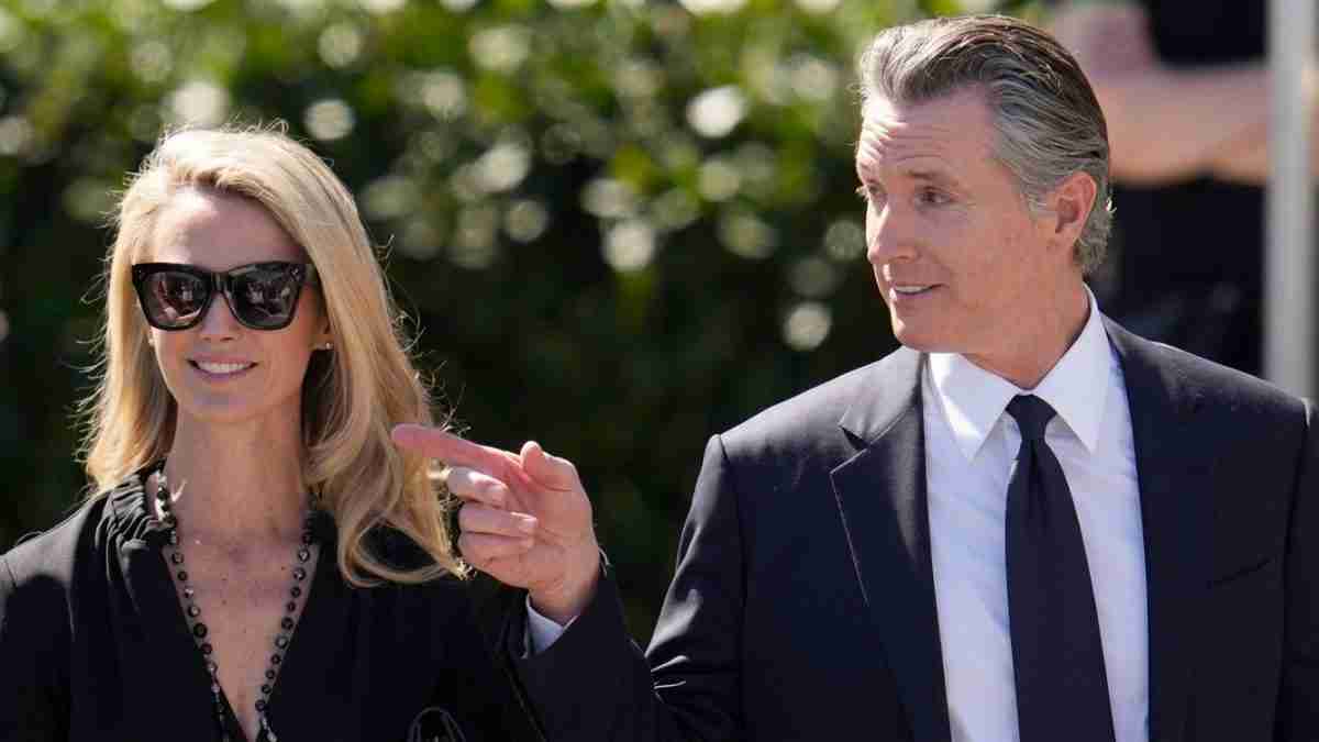 Man in a dark suit points at a smiling blonde woman wearing black sunglasses outdoors.