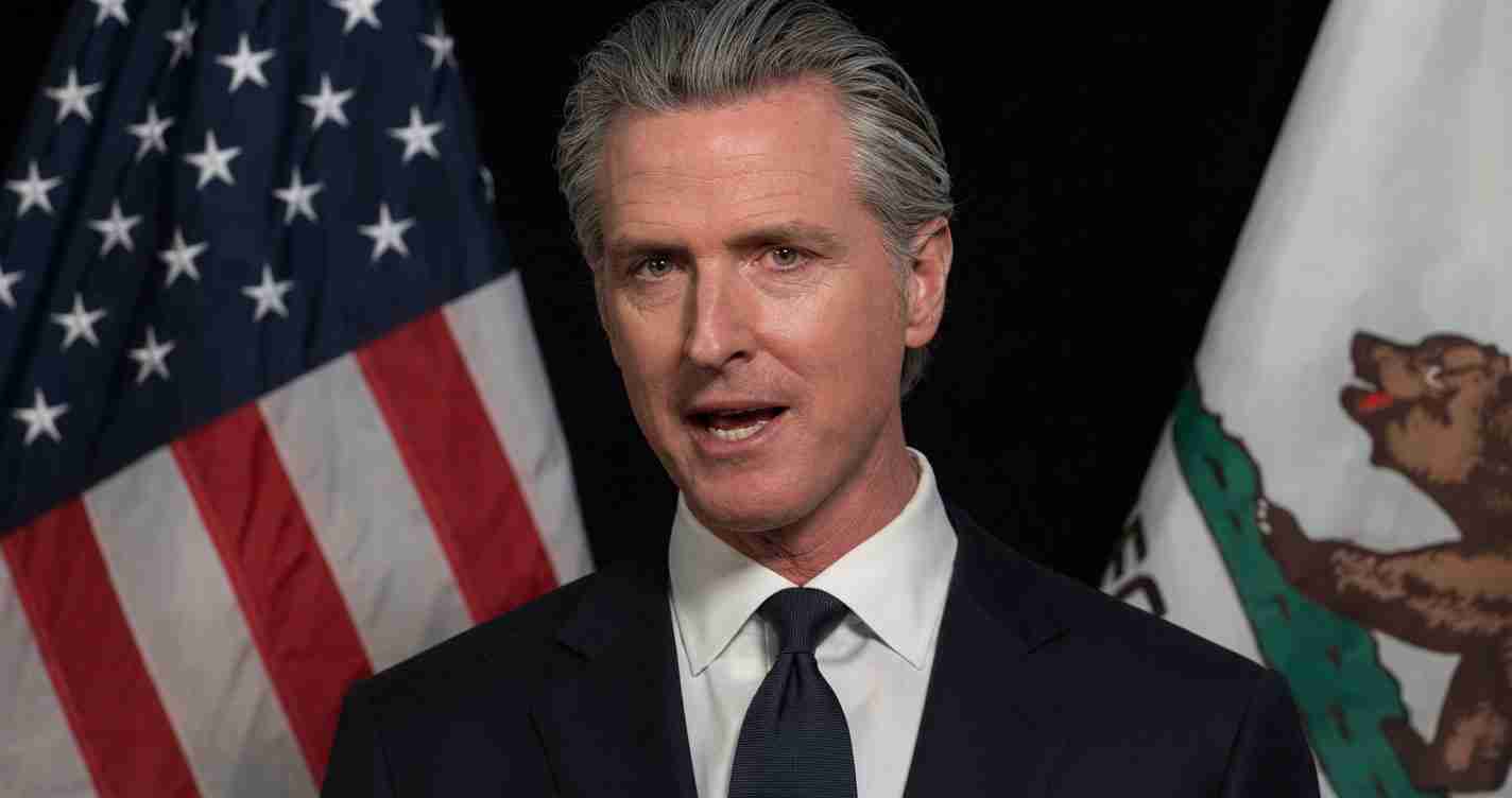 A suited man speaking at a formal event, with the American flag and a state flag in the background.