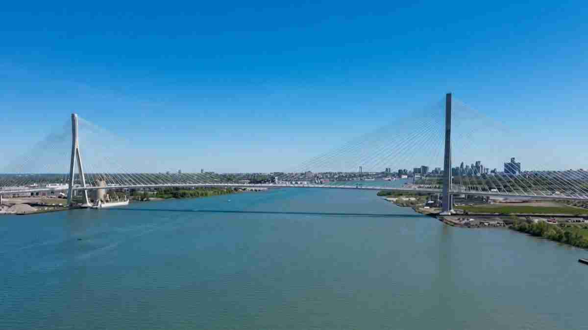 Cable-stayed bridge spanning a wide river with tall pylons and numerous cables, city skyline in the distance under a clear blue sky.