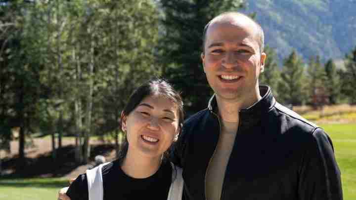 A smiling couple stands outdoors in a park with trees and mountains in the background, posing for a photo together.