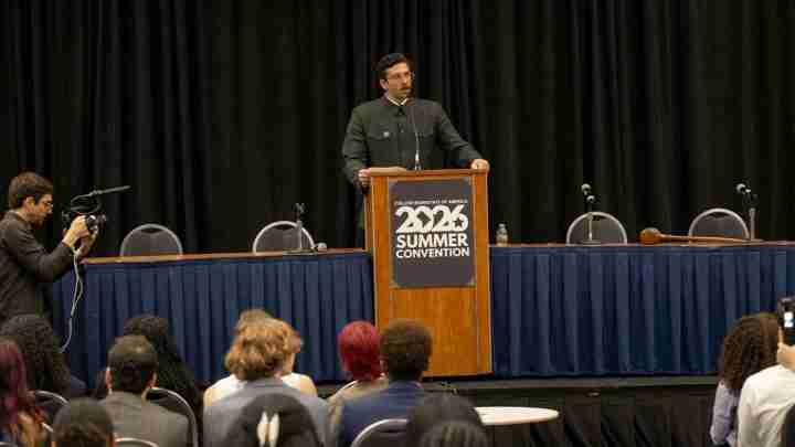 Speaker at a wooden podium delivering a talk on stage; photographer with camera films from the left, with an audience seated in front.