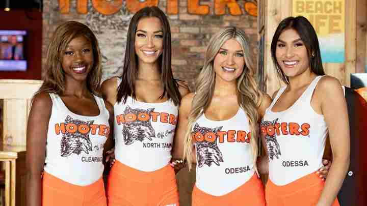 Four smiling Hooters waitresses in white tank tops with orange shorts pose together inside a rustic restaurant, ODESSA on two uniforms.