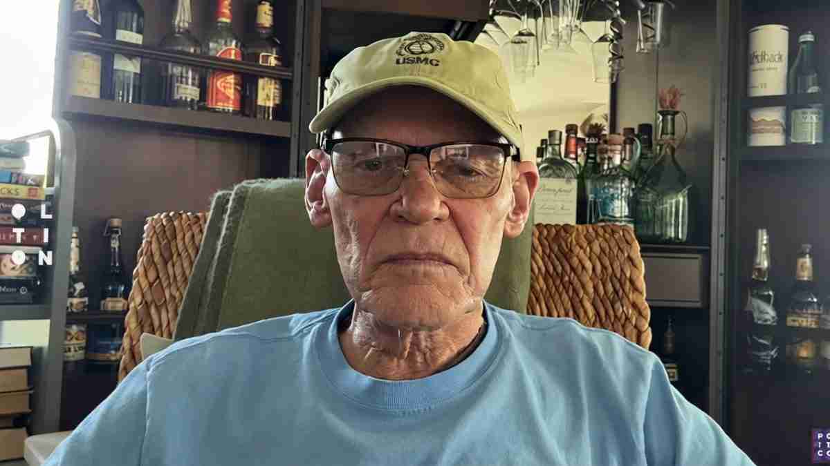 Senior man wearing a U.S. Marine cap and glasses sits facing the camera in a bar-like setting with shelves of bottles behind him.