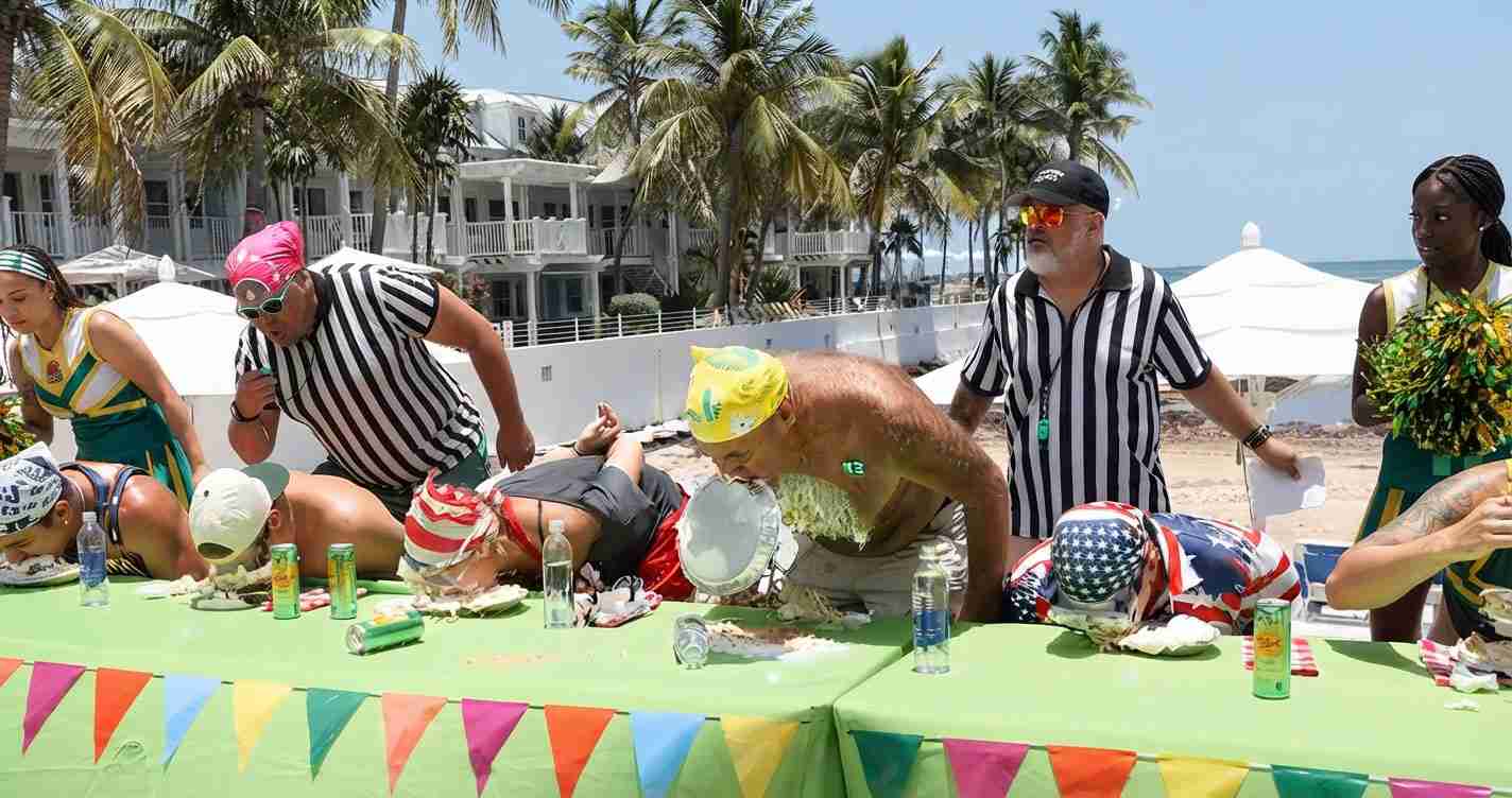 Group of people in swimwear and costumes take part in a beach-side eating contest at a green table under colorful pennant banners, with two referees in striped shirts and palm trees in the background.