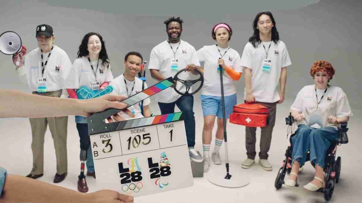 Clapperboard in the foreground as a diverse team of athletes and volunteers pose on a studio set with sports gear and assistive devices in the background.