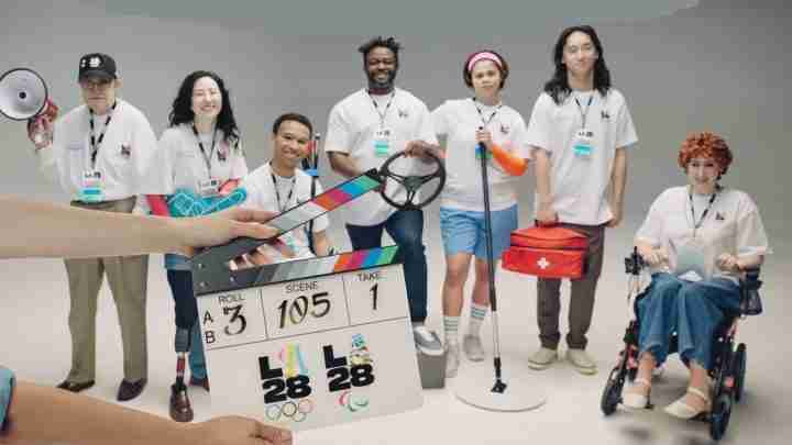 Clapperboard in the foreground as a diverse team of athletes and volunteers pose on a studio set with sports gear and assistive devices in the background.