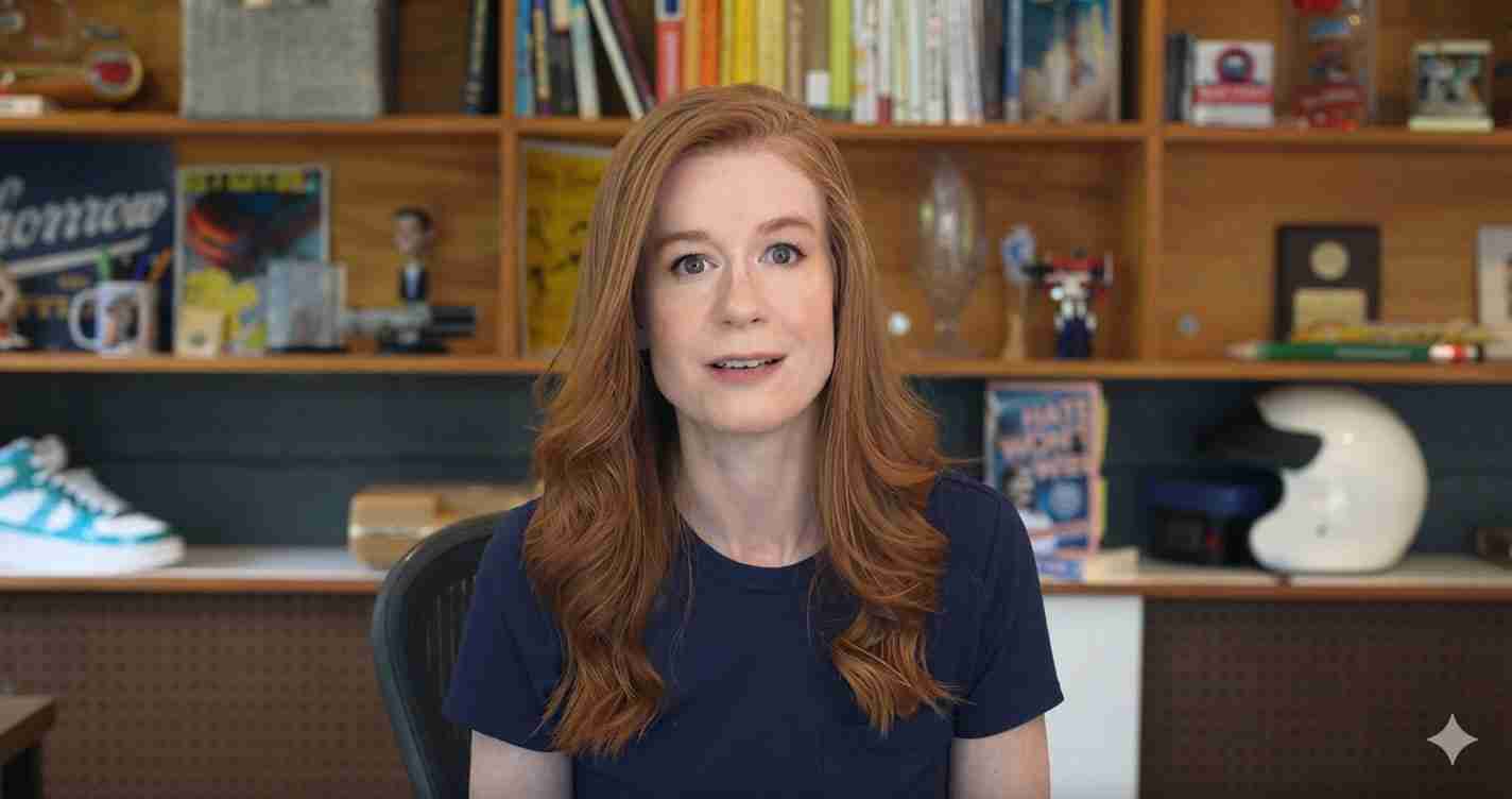 Red-haired woman in a navy shirt sits at a desk, facing the camera with a bookshelf-filled backdrop.