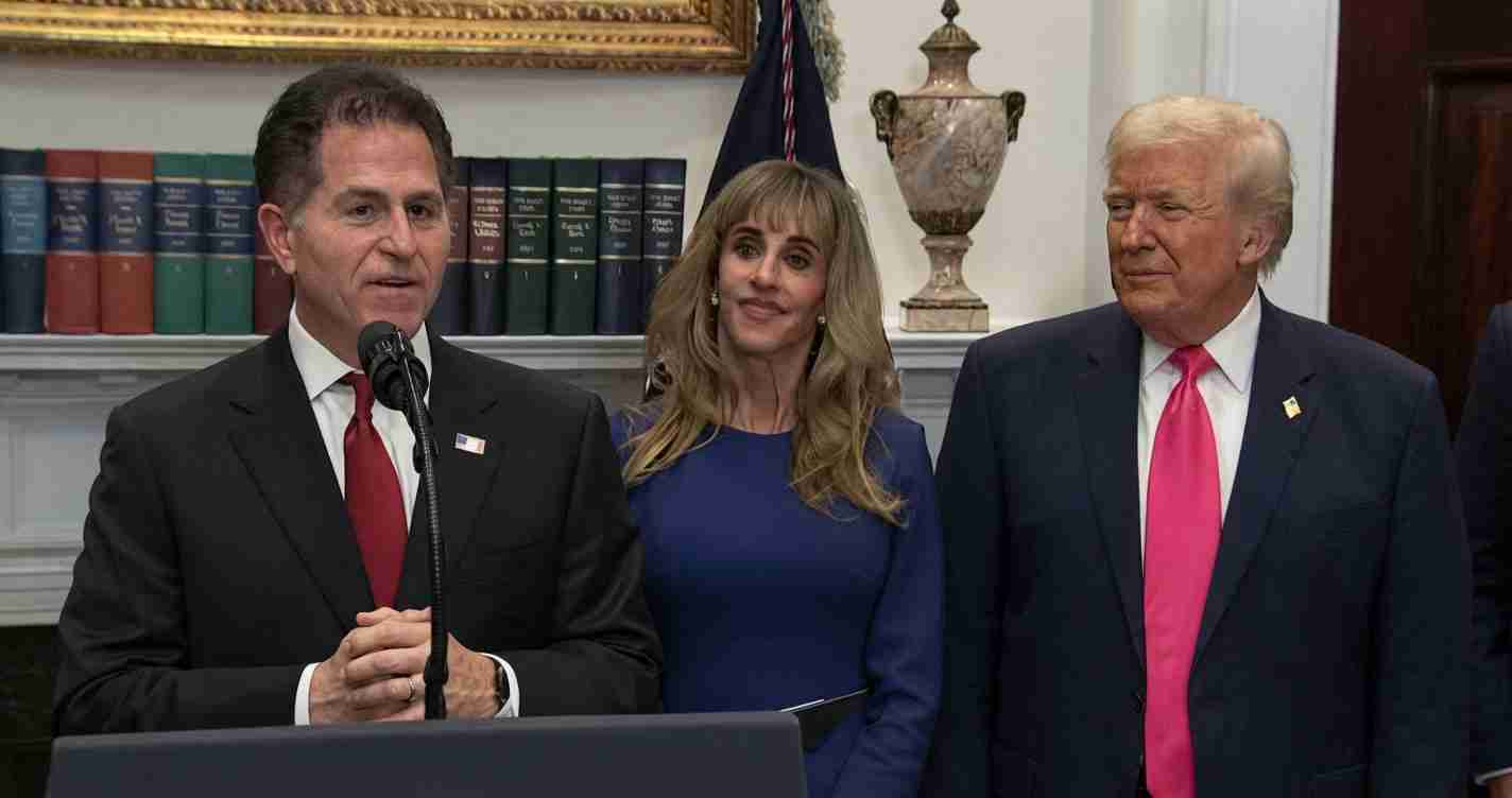 Man in a dark suit and red tie speaks at a podium with a microphone, flanked by a woman in blue and a man in a dark suit with a pink tie in a formal room with bookshelves and a decorative urn behind them.