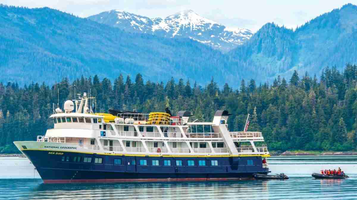 Large blue and white passenger cruise ship on calm water with forested mountains in the background, snow-capped peaks beyond the treeline.