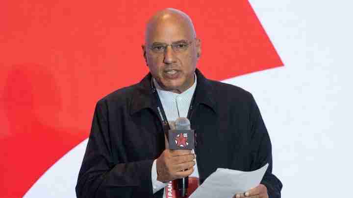 Bald man in a dark jacket speaks into a handheld microphone, holding a sheet of paper at a conference.