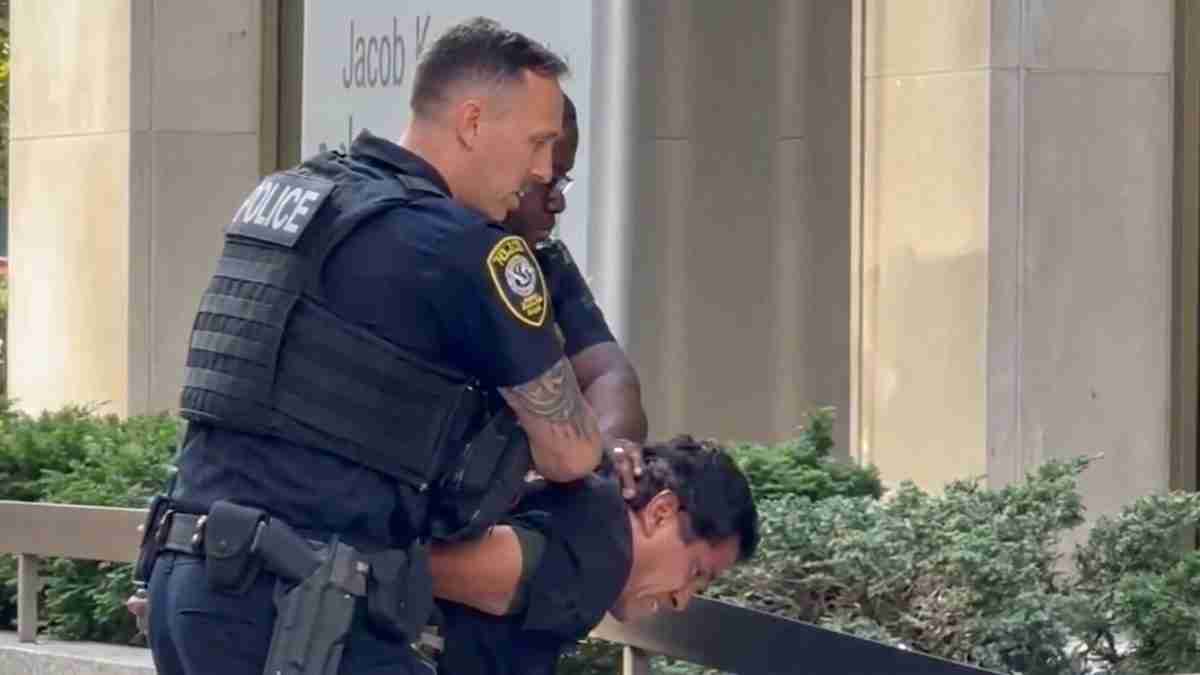 Two police officers restrain a man outside a building, one holding his arms behind his back.