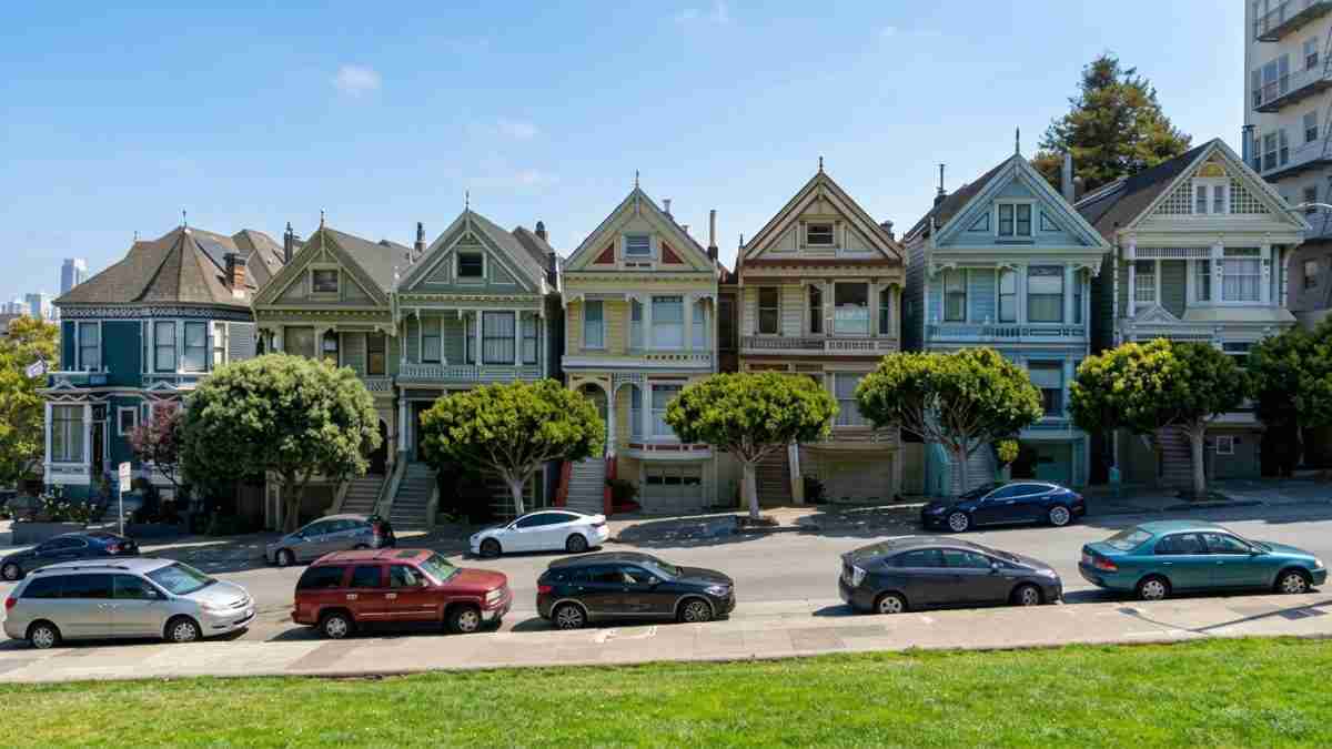 Row of colorful Victorian houses along a street with parked cars and trimmed trees in front on a sunny day, blue sky above.