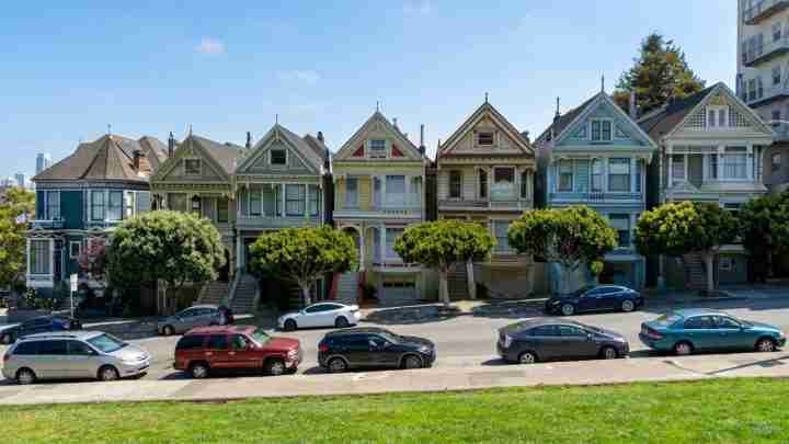 Row of colorful Victorian houses along a street with parked cars and trimmed trees in front on a sunny day, blue sky above.