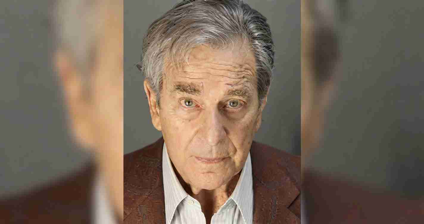 Close-up portrait of an older man with gray hair, wearing a brown blazer and light shirt, looking directly at the camera with a serious expression.