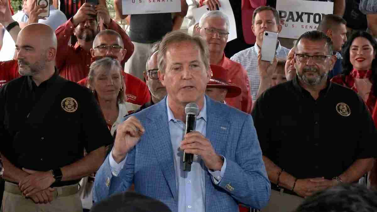 Man in a light blue blazer speaks into a handheld microphone at a crowded rally, supporters behind him holding signs and cameras.