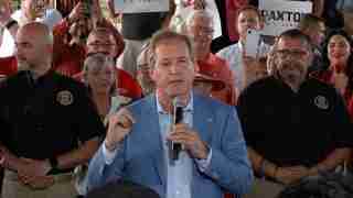 Man in a light blue blazer speaks into a handheld microphone at a crowded rally, supporters behind him holding signs and cameras.