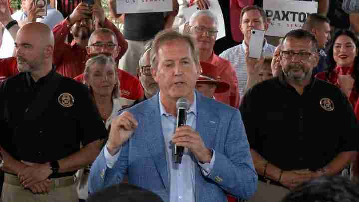 Man in a light blue blazer speaks into a handheld microphone at a crowded rally, supporters behind him holding signs and cameras.