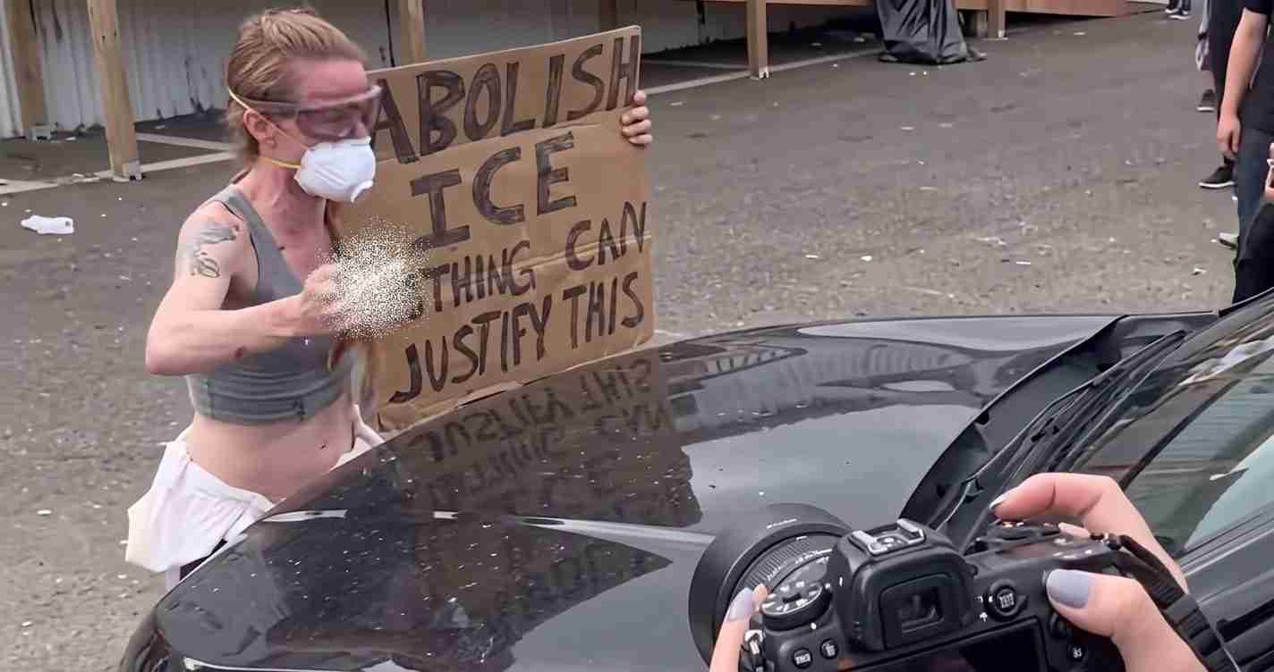 Masked protester in goggles and a gray sports bra holds a cardboard sign that reads 'ABOLISH ICE' beside a dark car, as a photographer shoots.