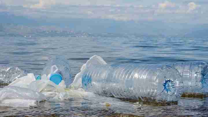 Floating plastic bottles and bags drift on the calm sea near the shore, highlighting pollution, with distant mountains in the background.