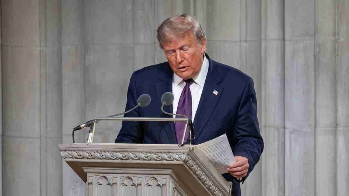 Man in a navy suit and purple tie speaks at a stone podium, reading from a document with two microphones in front.