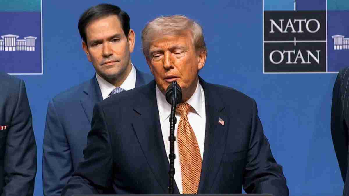 Man in a dark suit and orange tie speaks at a podium with a NATO backdrop; two men stand behind him.