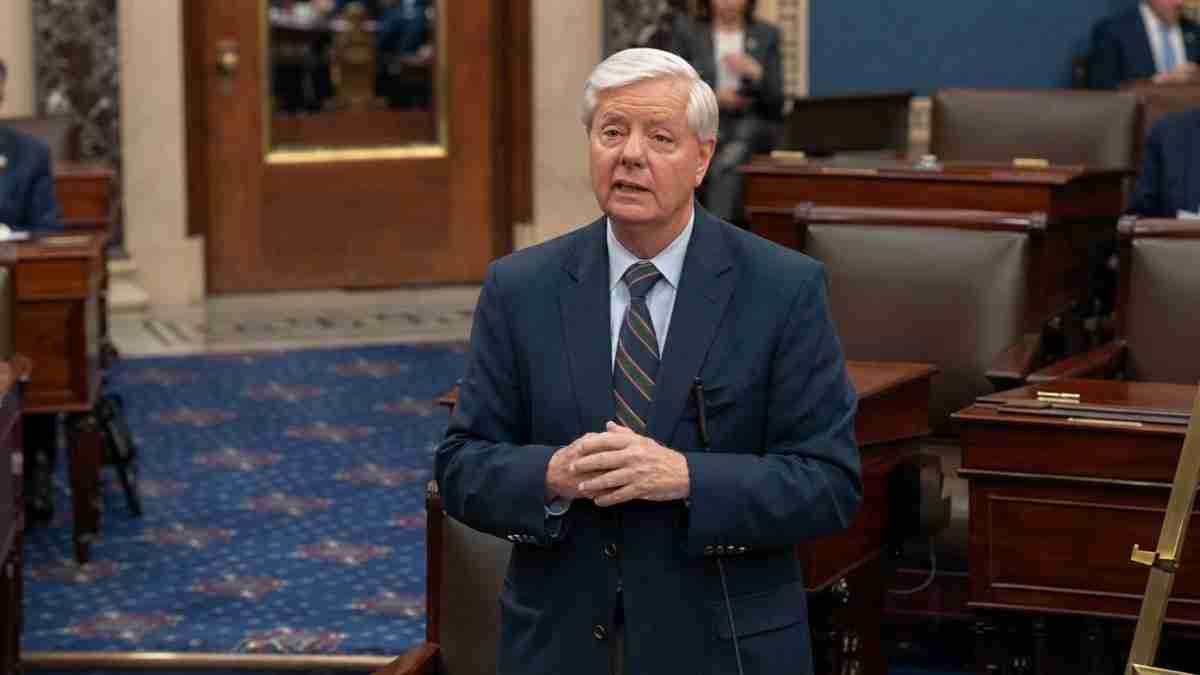 A suited man with white hair speaks to colleagues in a formal legislative chamber, hands clasped in front of him.
