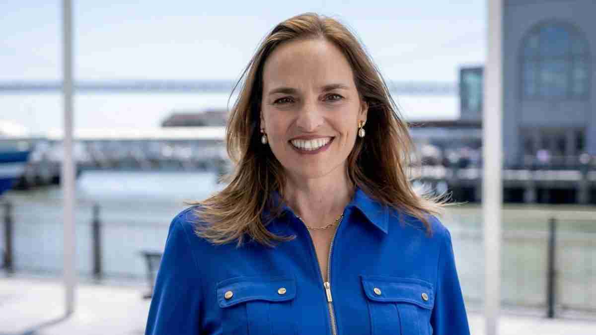 Smiling woman in a blue button-down shirt stands by a marina with boats in the background.