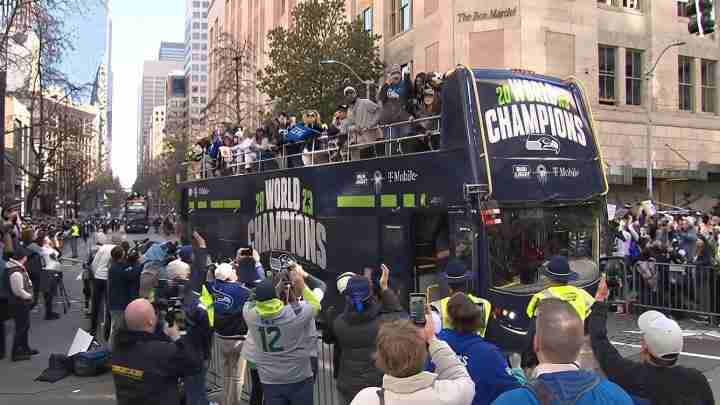 Double-decker bus in navy with 'World Champions' and Seahawks logos; fans cheer from the open top deck as a city parade passes with crowds along barricades.