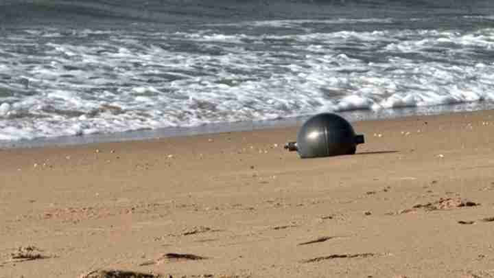 Large black spherical buoy washed up on a sandy beach with gentle waves in the background and sea foam nearby.