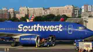 Blue Southwest Airlines jet at a gate, with a jet bridge attached and ground crew preparing the aircraft; 'Southwest' logo across the fuselage and 'Southwest.com' on the engine nacelle present in the foreground.