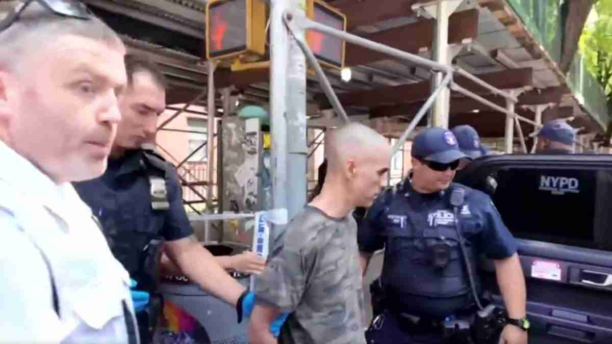 A man in a gray camouflage shirt is being handcuffed by two police officers beside a NYPD vehicle, with scaffolding and a construction site in the background.