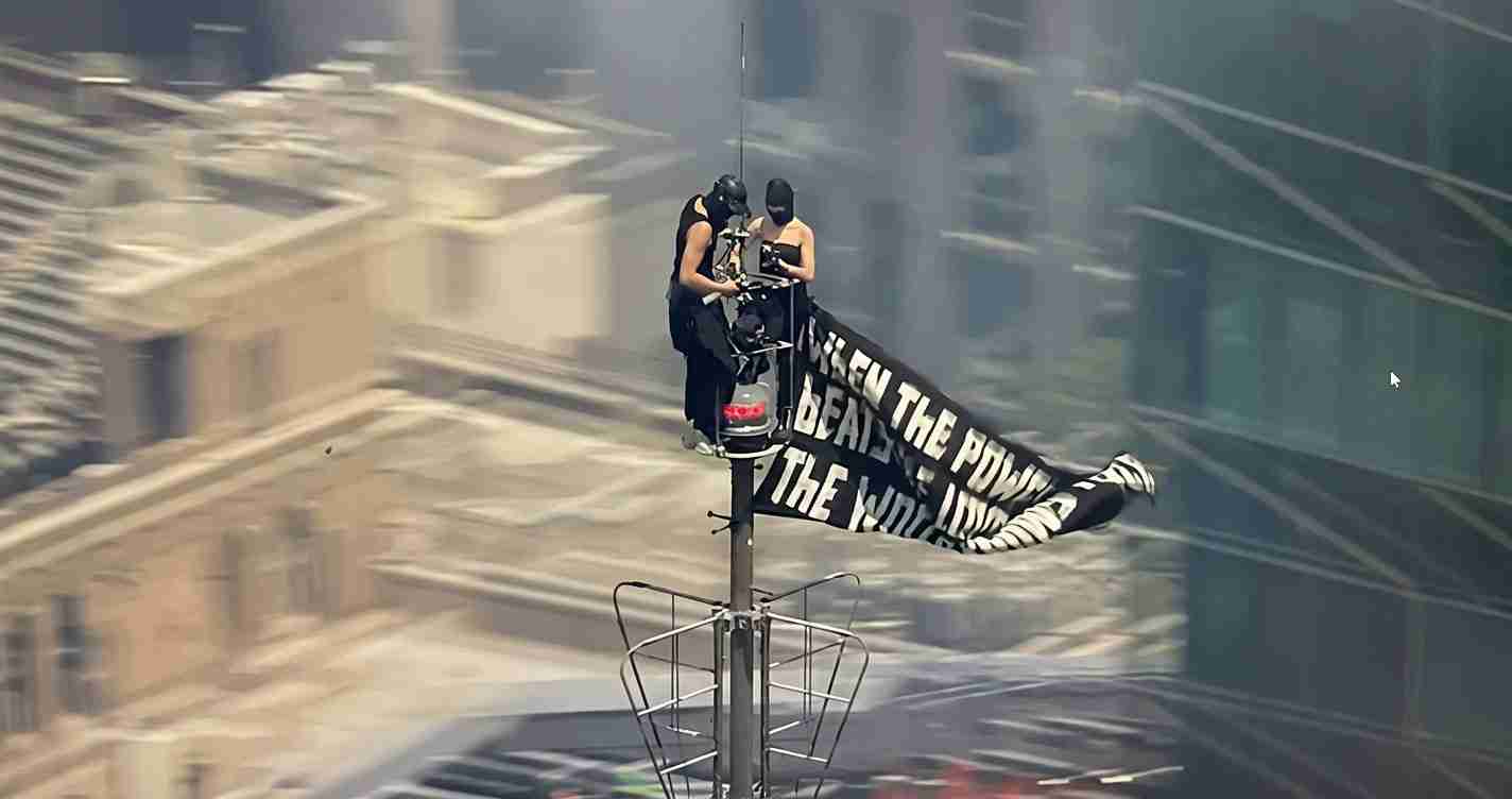 Two people in black outfits and helmets stand atop a pole on a city rooftop, unfurling a long black banner with large white text over a blurred urban street below.