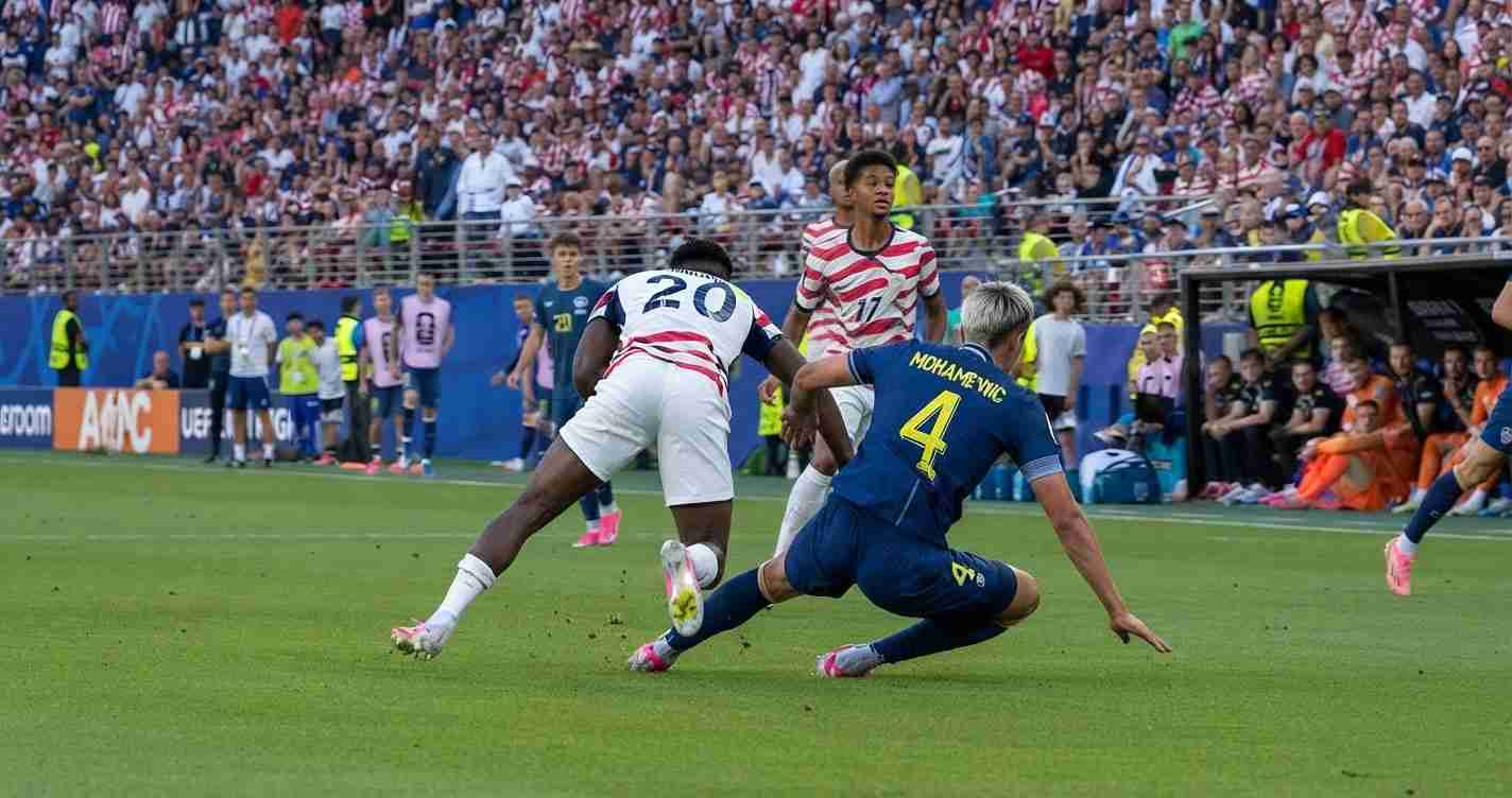Soccer match moment: a player in white and navy tackles the ball while a blue jersey defender slides in from the right, crowd in the stands nearby.