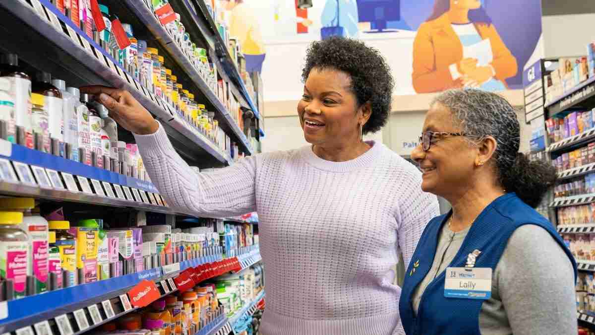 Two women in a pharmacy aisle, one shopper selecting a bottle from the shelf while the other, a store employee in a blue vest with a name badge, smiles and observes.